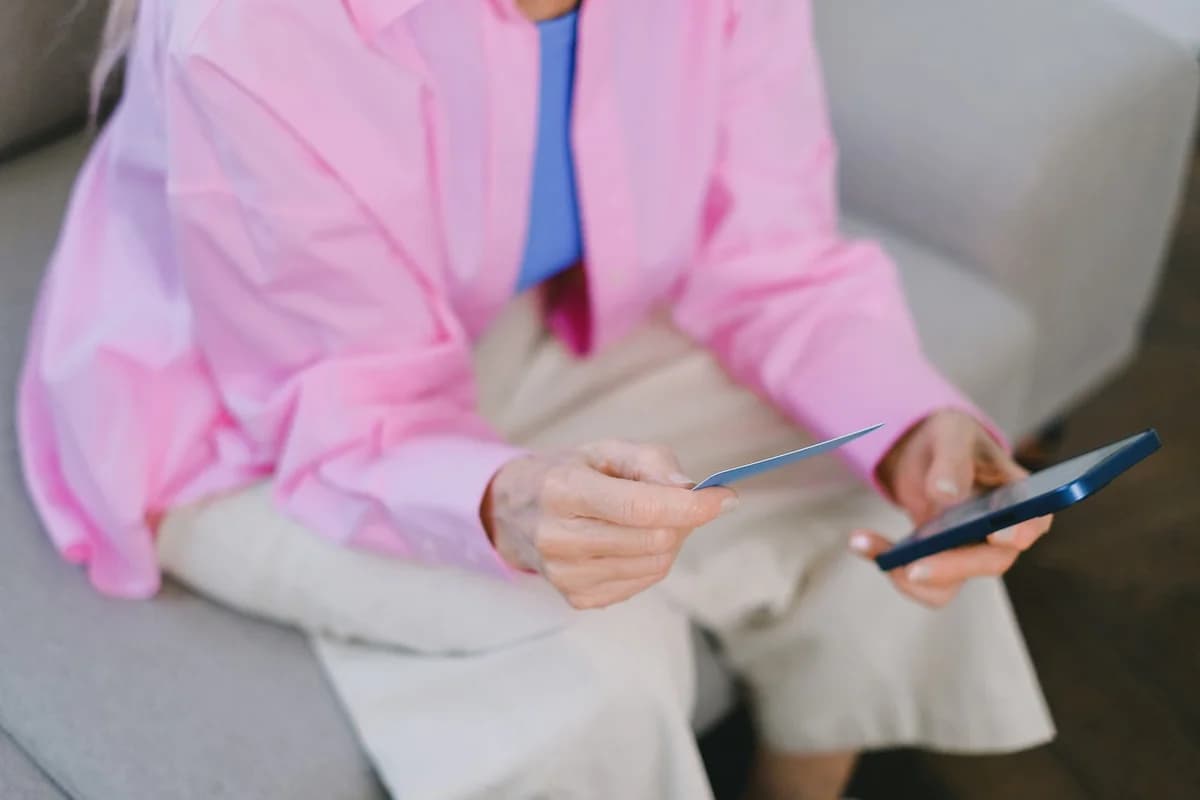 An older woman holds a credit card and smartphone while sitting on a couch.