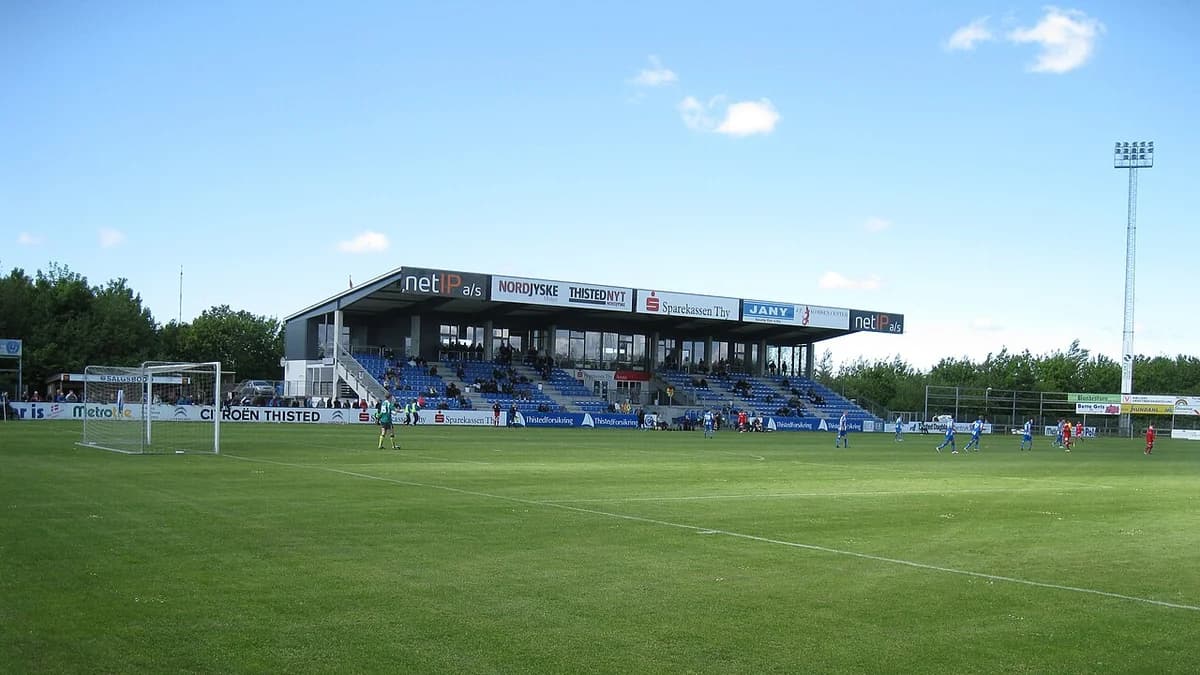 Wide view of a football stadium in Thisted with stands and players on the pitch.