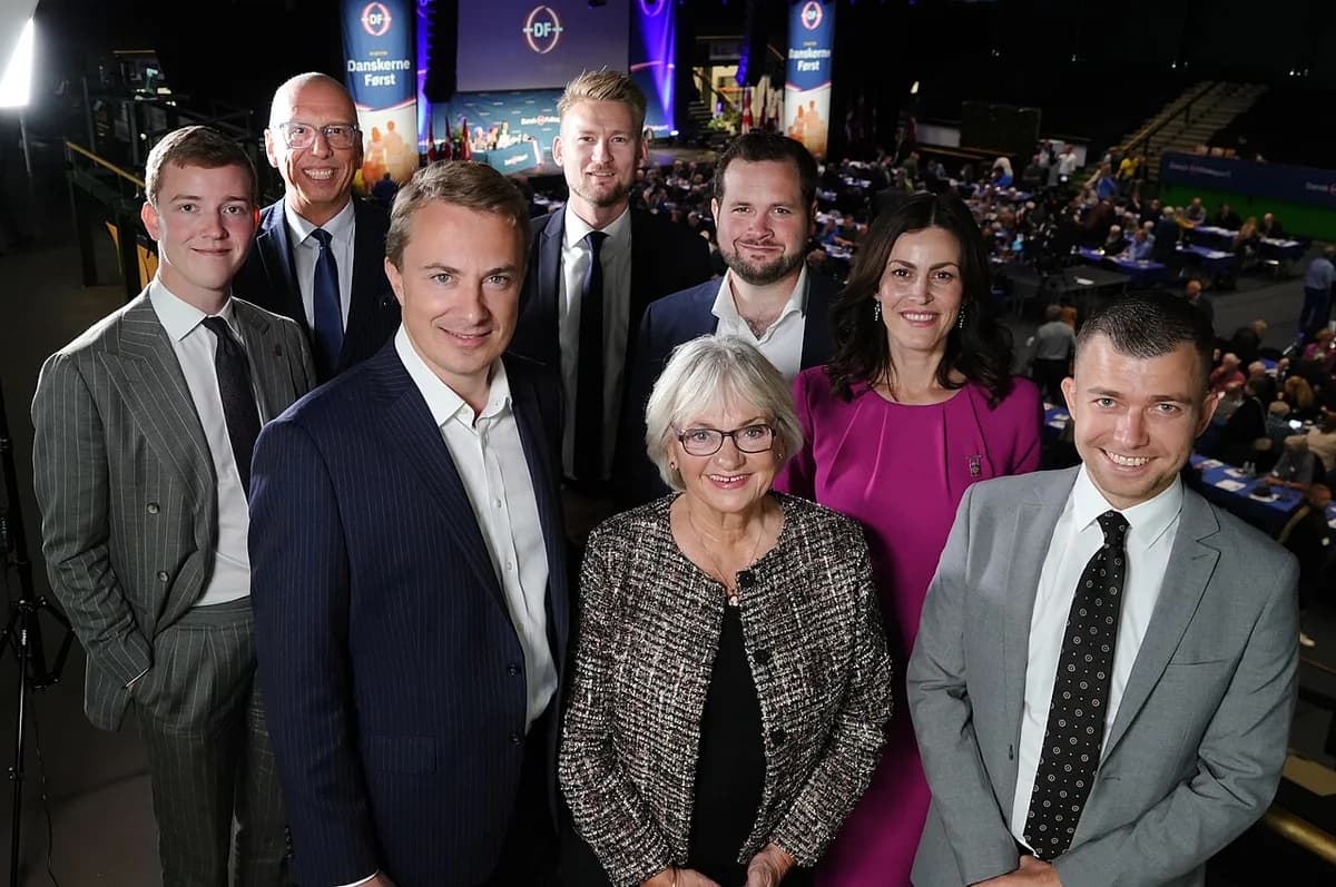 Seven Danish politicians posing at an indoor party meeting with an audience in the background.