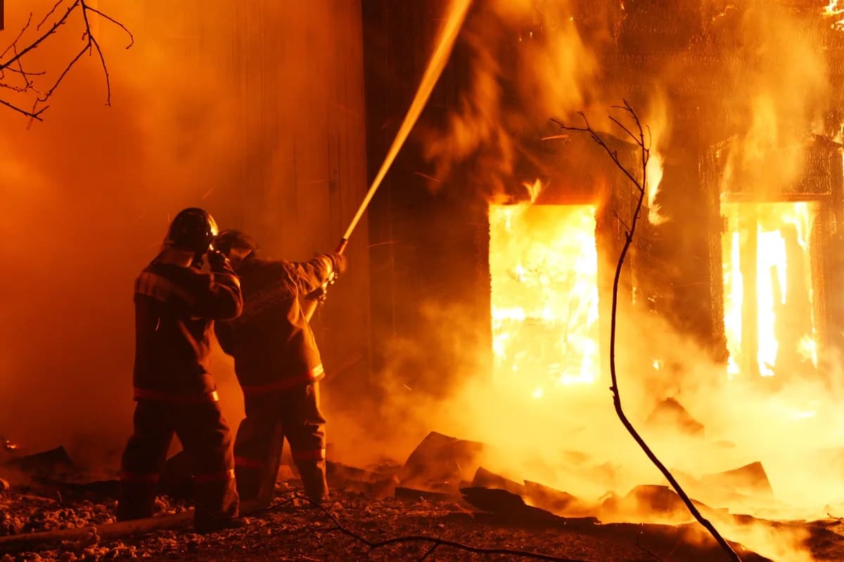 Two firefighters spray water at a burning building at night.
