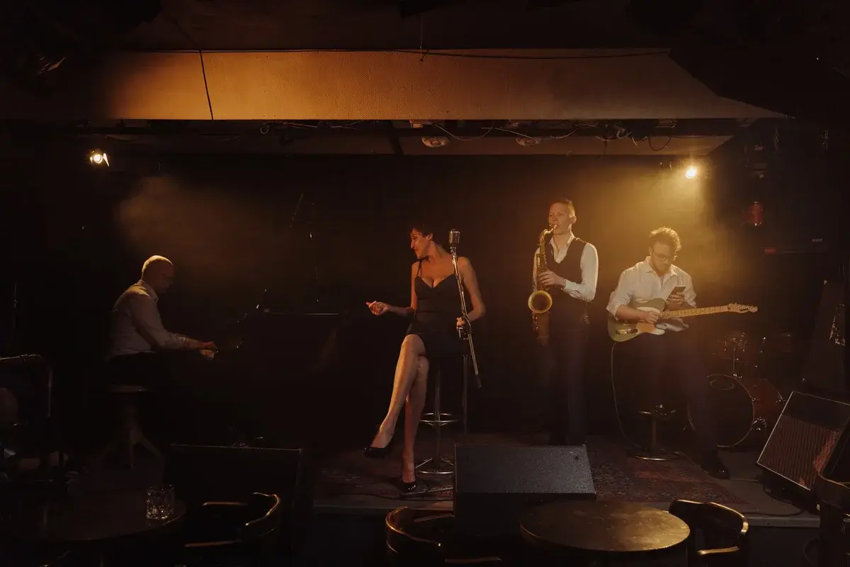 A jazz band performs on a dimly lit stage with a female singer, pianist, saxophonist, and guitarist.