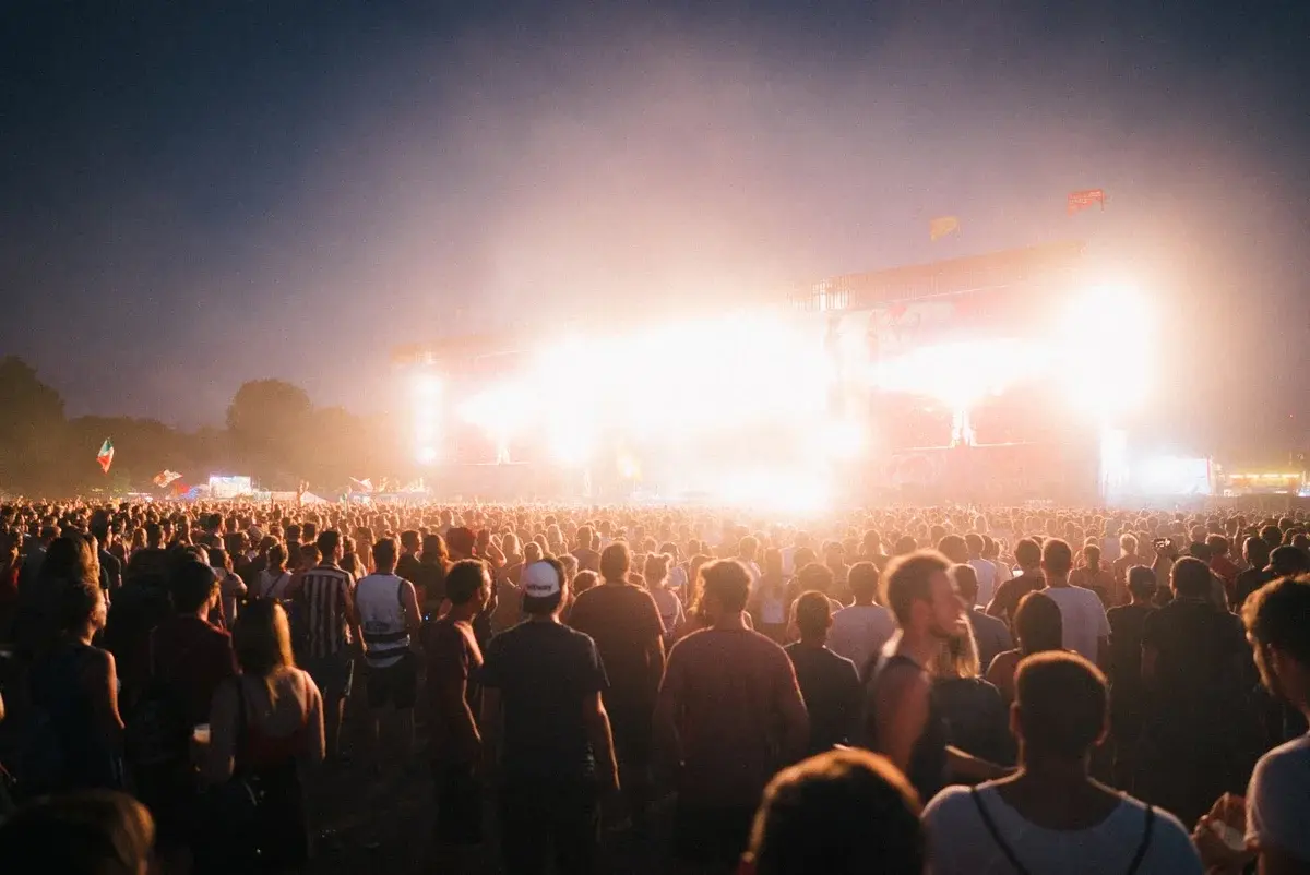 A large nighttime crowd watches a brightly lit outdoor concert stage.