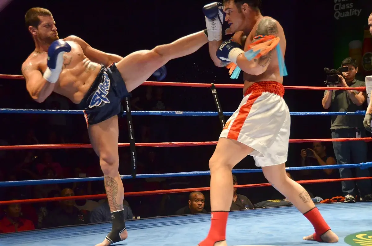 Two Muay Thai fighters; one delivers a high kick while the other blocks it.