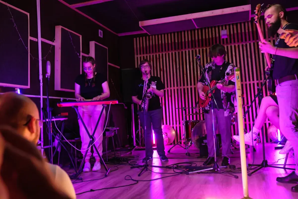 A small band performs indoors under pink-purple stage lighting.
