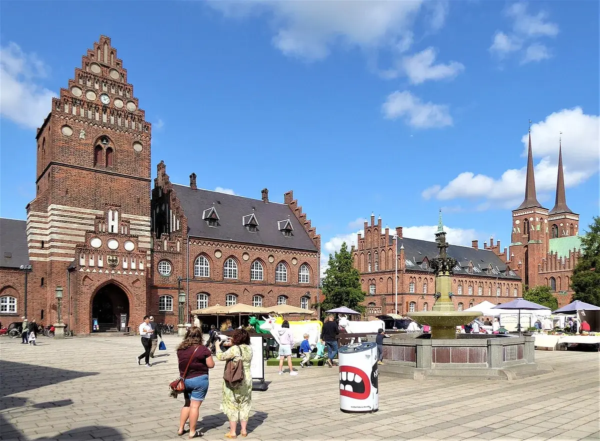 Historic red-brick town hall square in Roskilde with a fountain, market stalls, and church spires under a blue sky.