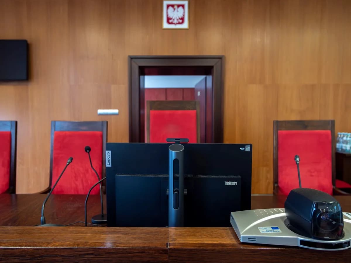 Modern courtroom with red chairs, microphones, a computer monitor, and the Polish coat of arms above the bench.