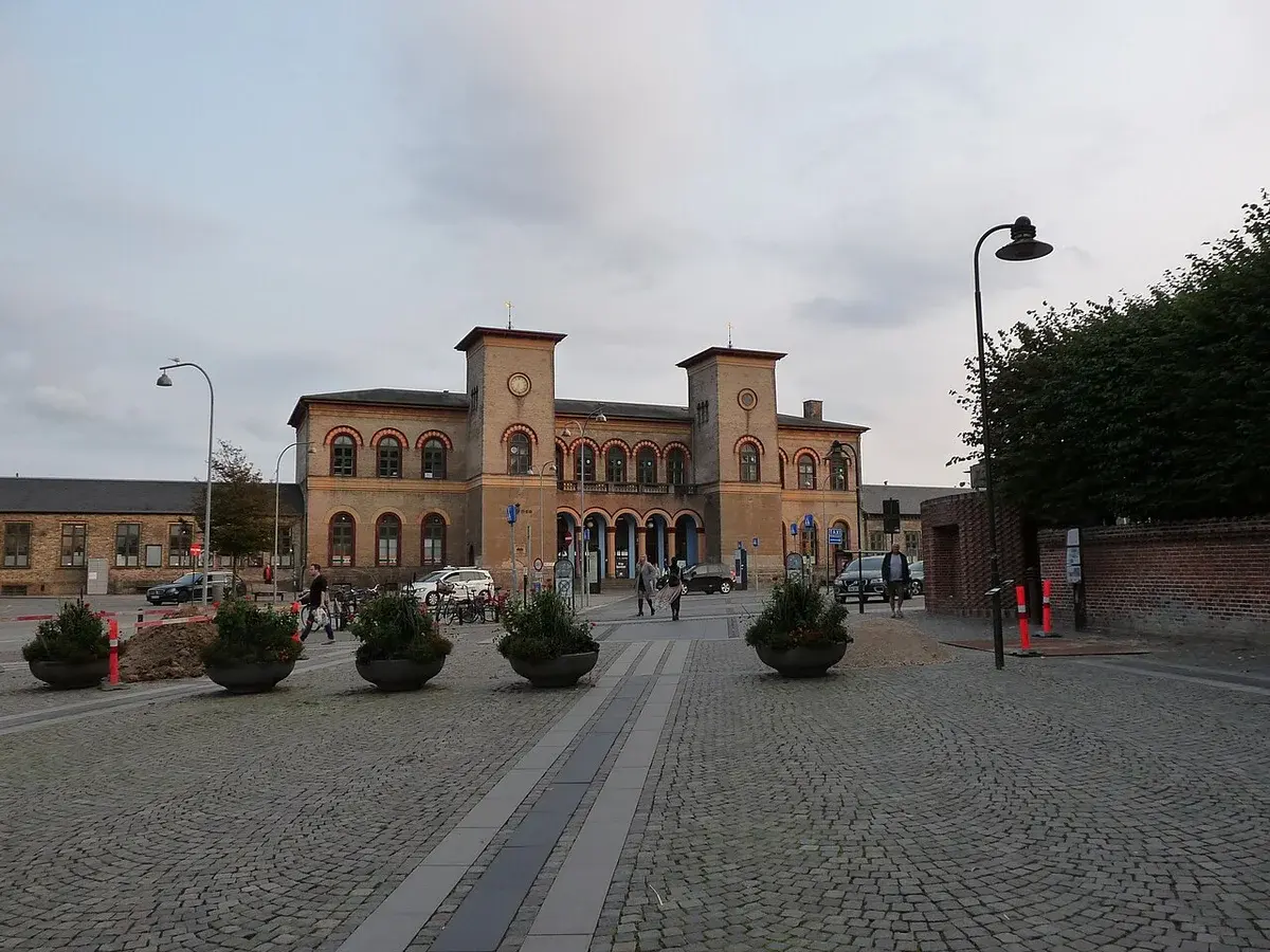 Exterior view of Roskilde Station with a cobblestone forecourt and people walking nearby.