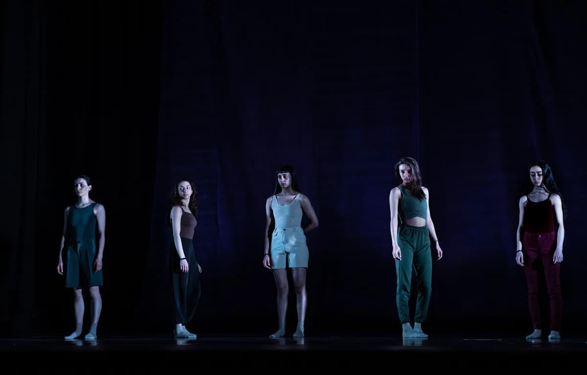 Five female dancers standing on a dark stage.