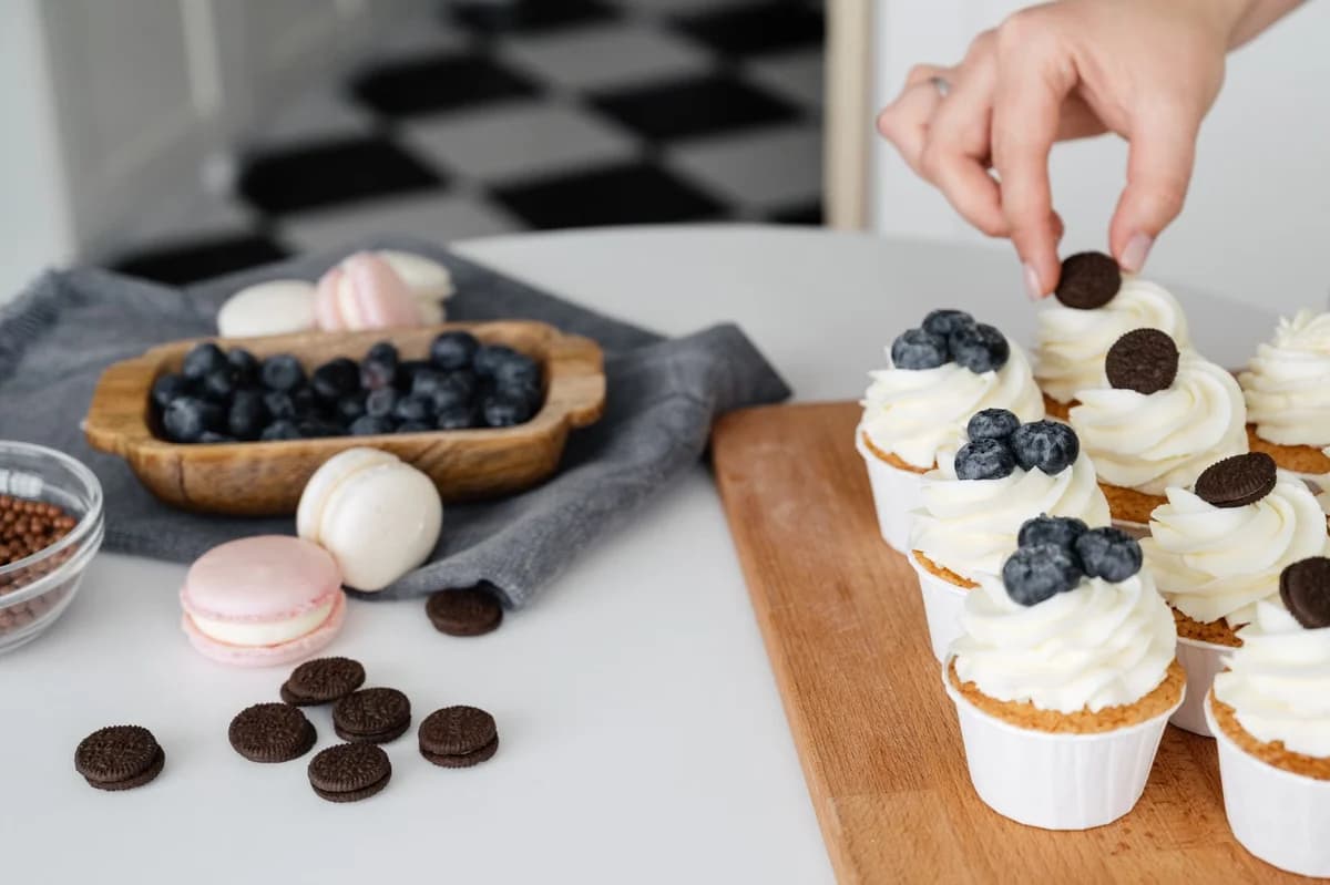 A hand decorating frosted cupcakes with blueberries and chocolate cookies on a table.