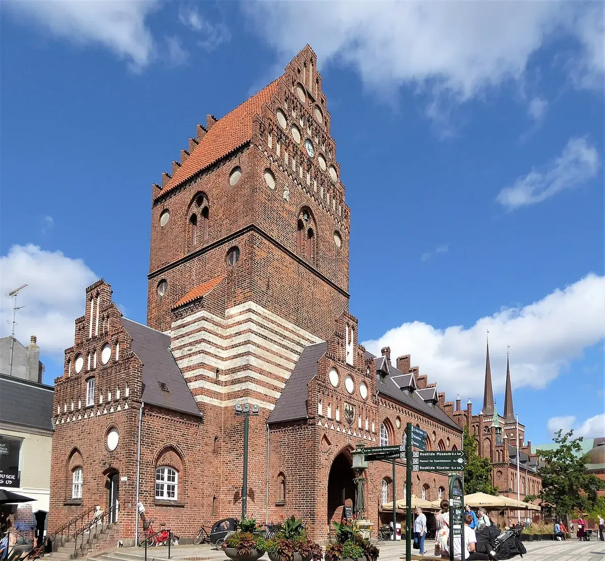 Historic brick town hall in Roskilde on a sunny day with people, bicycles, and street signs.
