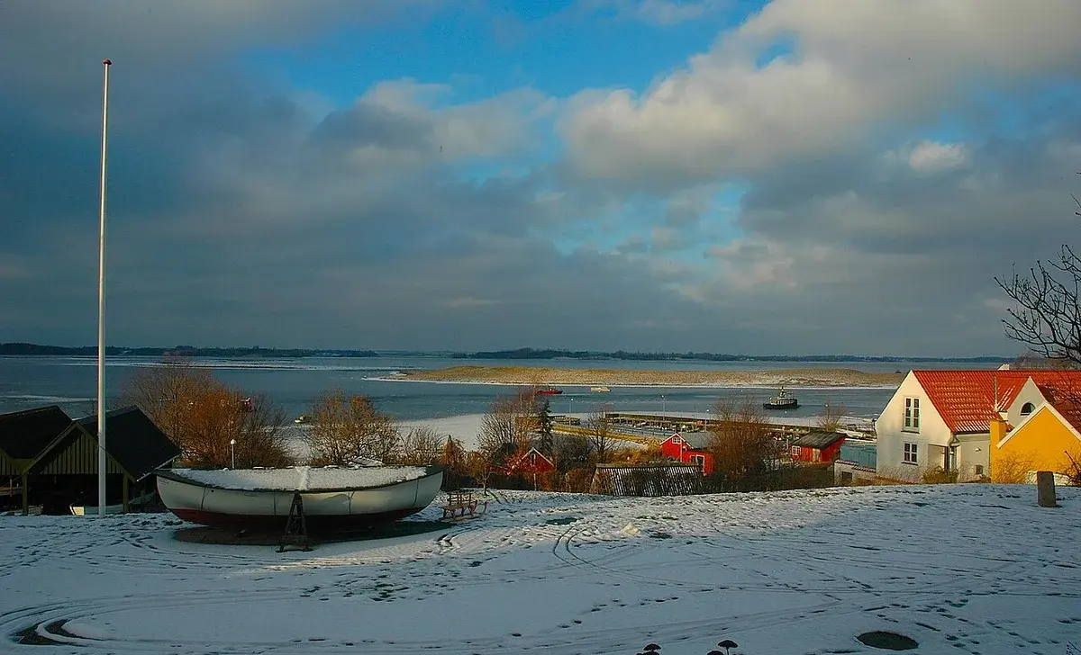Snowy winter view over Roskilde Fjord with an upside-down boat and colorful houses on the shore.