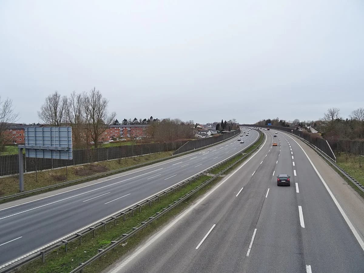 Light traffic on a Danish motorway under a cloudy sky, with houses and trees along the sides.